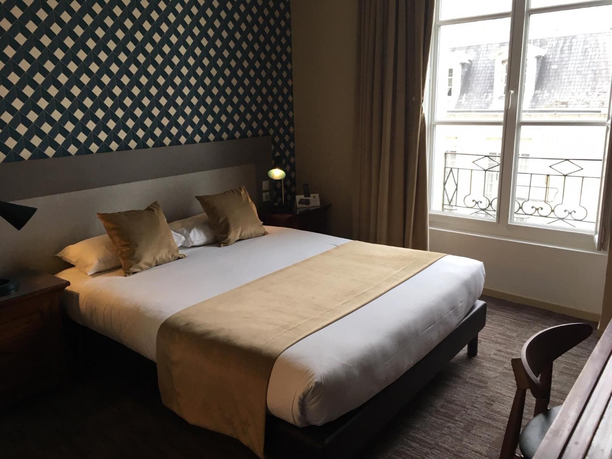 A neatly made bed with a headboard, a bedside lamp, and a view of the street through a window with a metal railing.