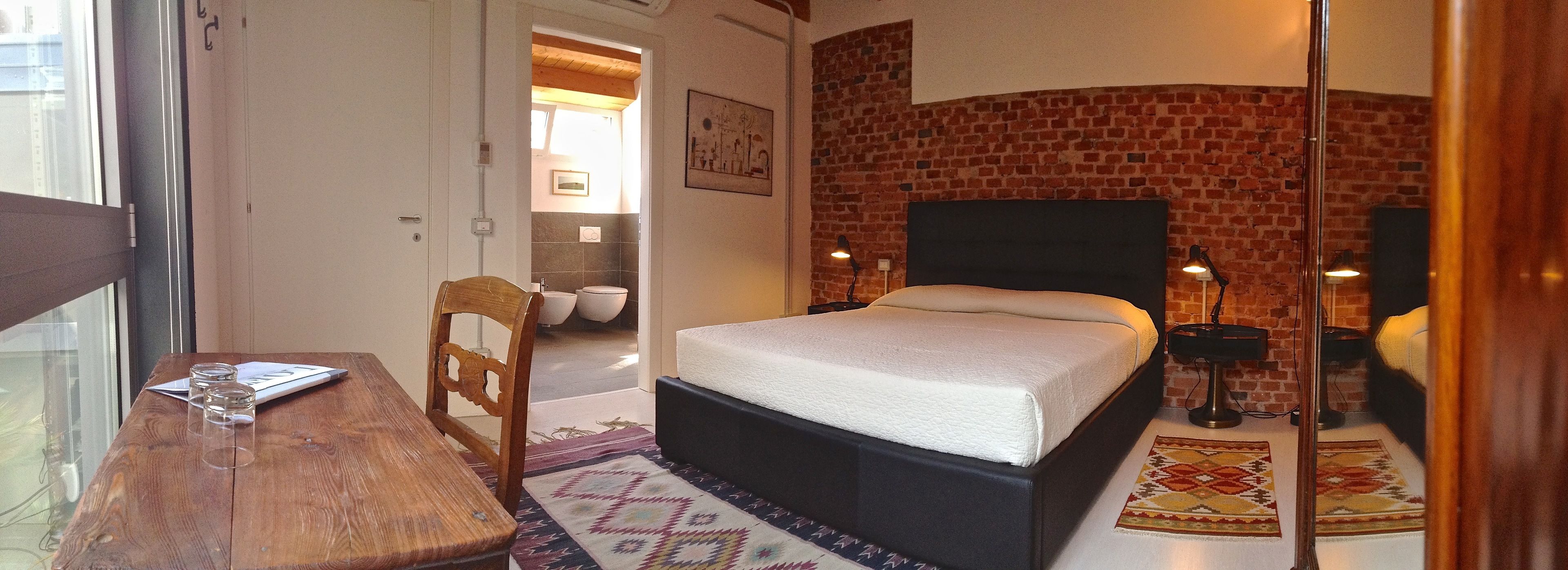A bedroom with a brick wall, a bed, a wooden chair, a table with glasses, and a rug.