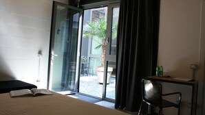 A hotel room with a bed, a desk, a chair, and a sliding glass door leading to a courtyard with a palm tree and a potted plant. - LoftVerona (Verona)