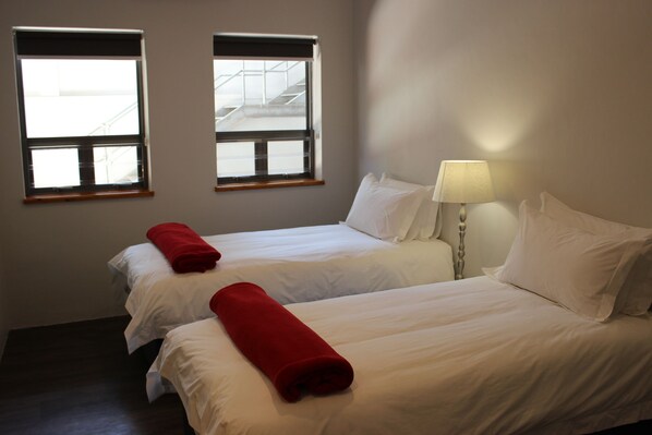A hotel room with two beds, each with white linens and a red towel. There is a window on the left and a lamp on the right. - Zeederberg Corner (Paarl)