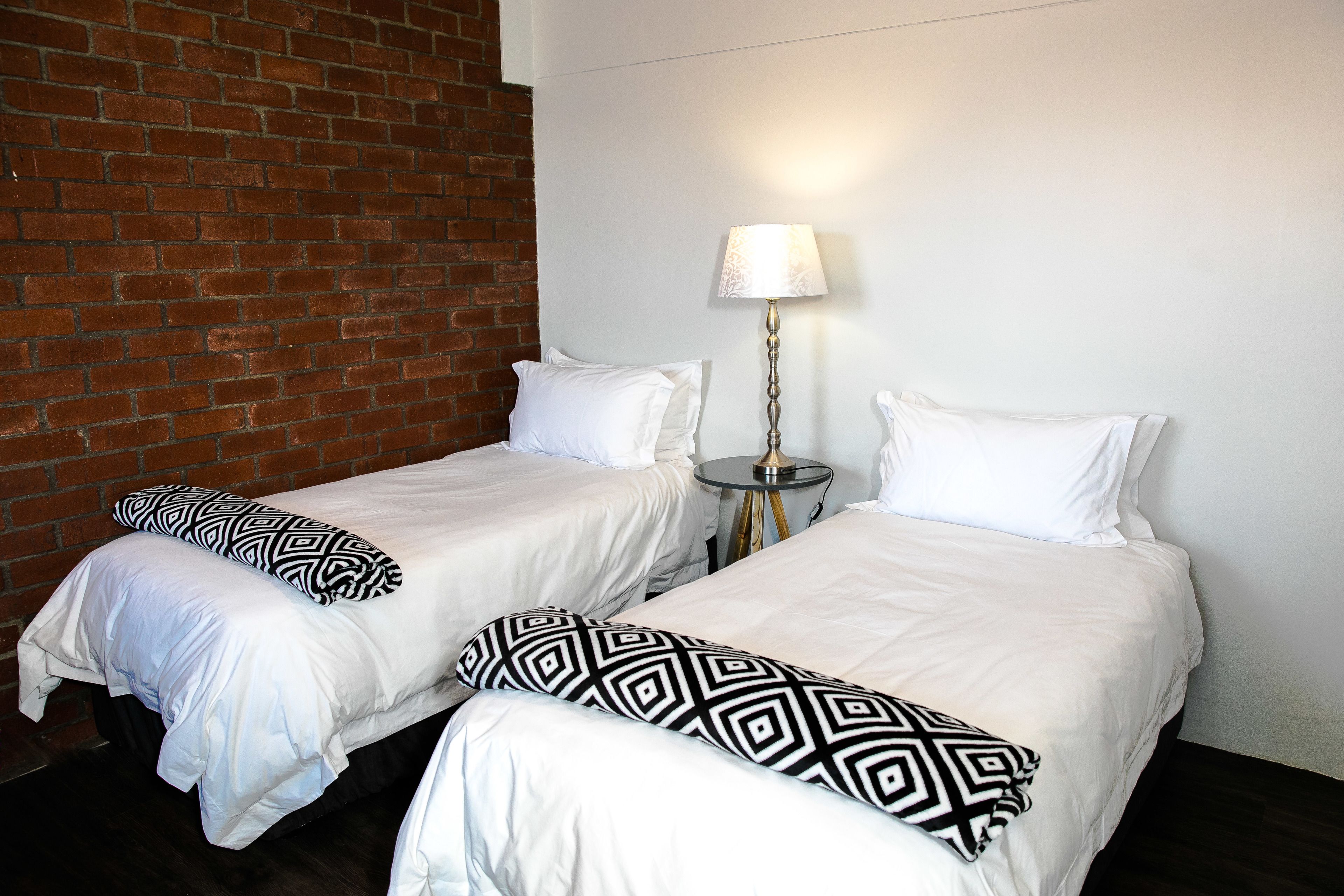 Two single beds with white bedding and black geometric pillows, a small round table with a lamp, and a brick wall.