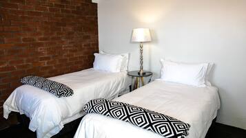 Two single beds with white bedding and black geometric pillows, a small round table with a lamp, and a brick wall.
