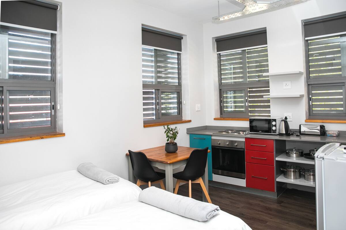 Habitación compacta y moderna con cama, una pequeña mesa de comedor con sillas, kitchenette y ventanas amplias con persianas.