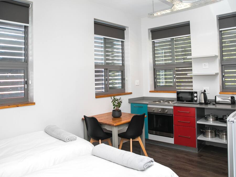 A compact, modern room with a bed, a small dining table with chairs, a kitchenette, and large windows with blinds.