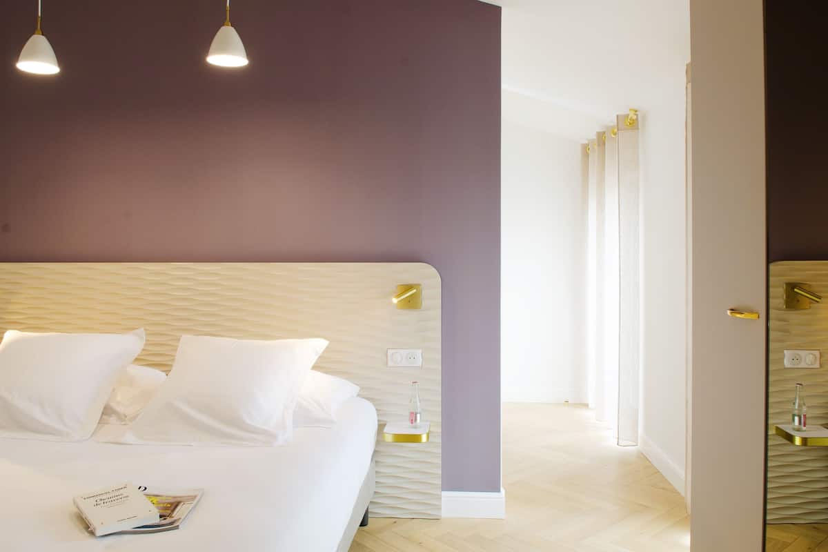 A modern bedroom with a large bed, white bedding, and a magazine on the bed. There are two pendant lights above the bed, and the room has a wooden floor and a purple accent wall.