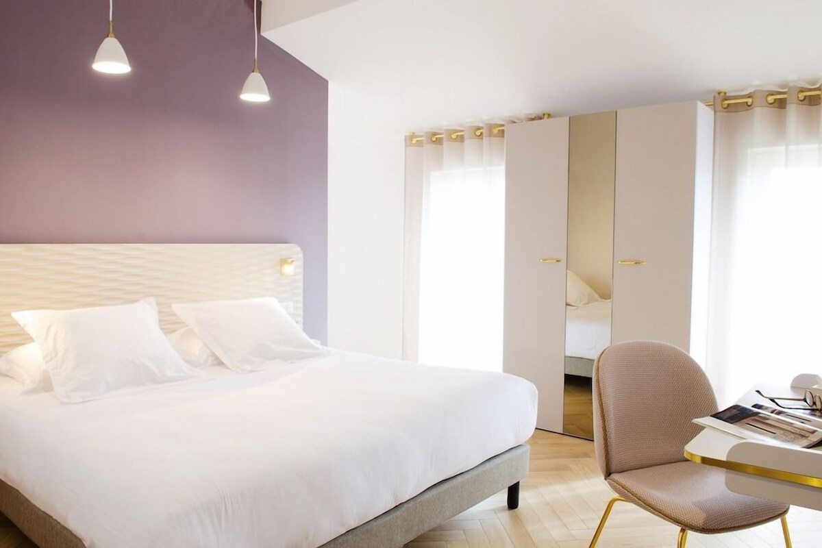A modern hotel room with a large bed, a desk with a chair, and a wardrobe with a mirror.