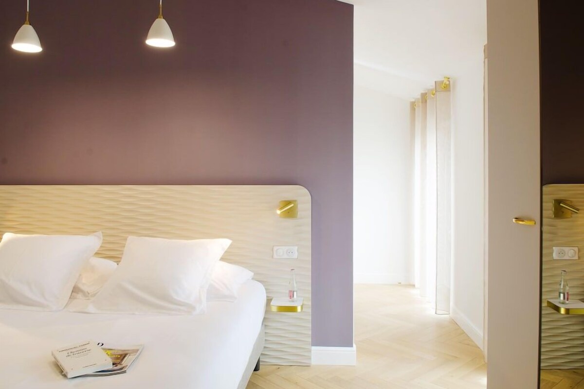 A modern hotel room with a large bed, white linens, a magazine on the bed, pendant lights, and a view of the hallway.