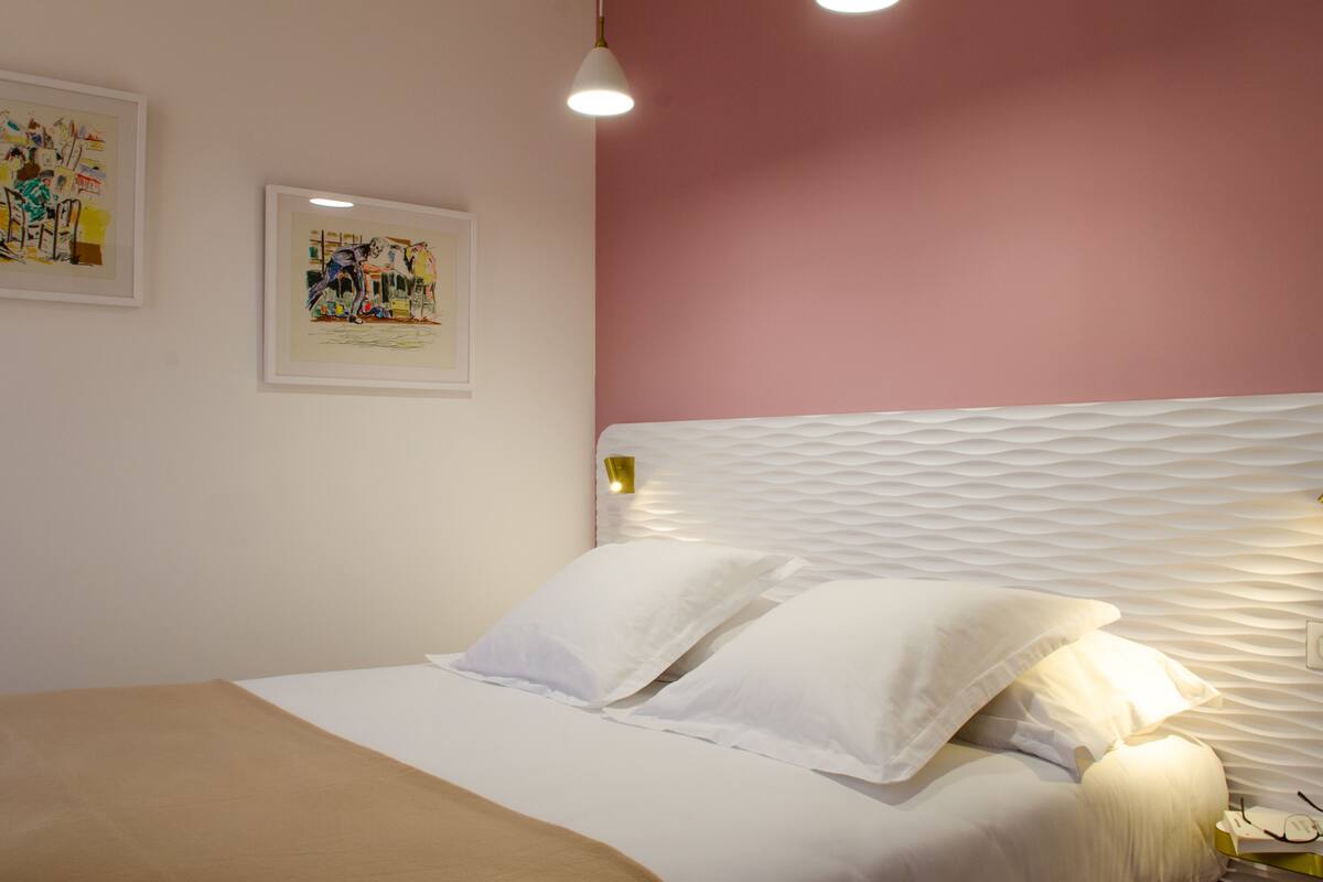 A neatly made bed with white linens and pillows, a bedside table with a lamp, and two framed artworks on the wall.