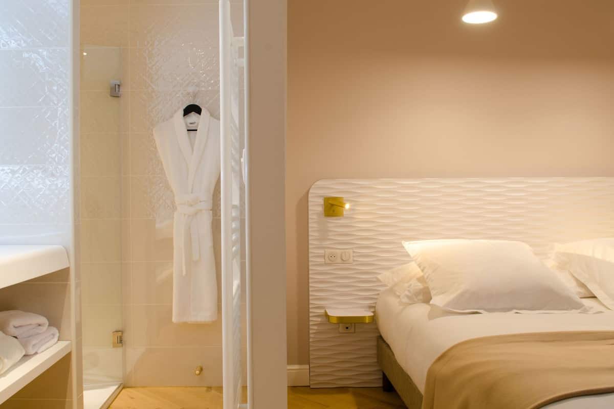 A hotel room with a shower, a bed with a textured headboard, and a white robe hanging on a rack.
