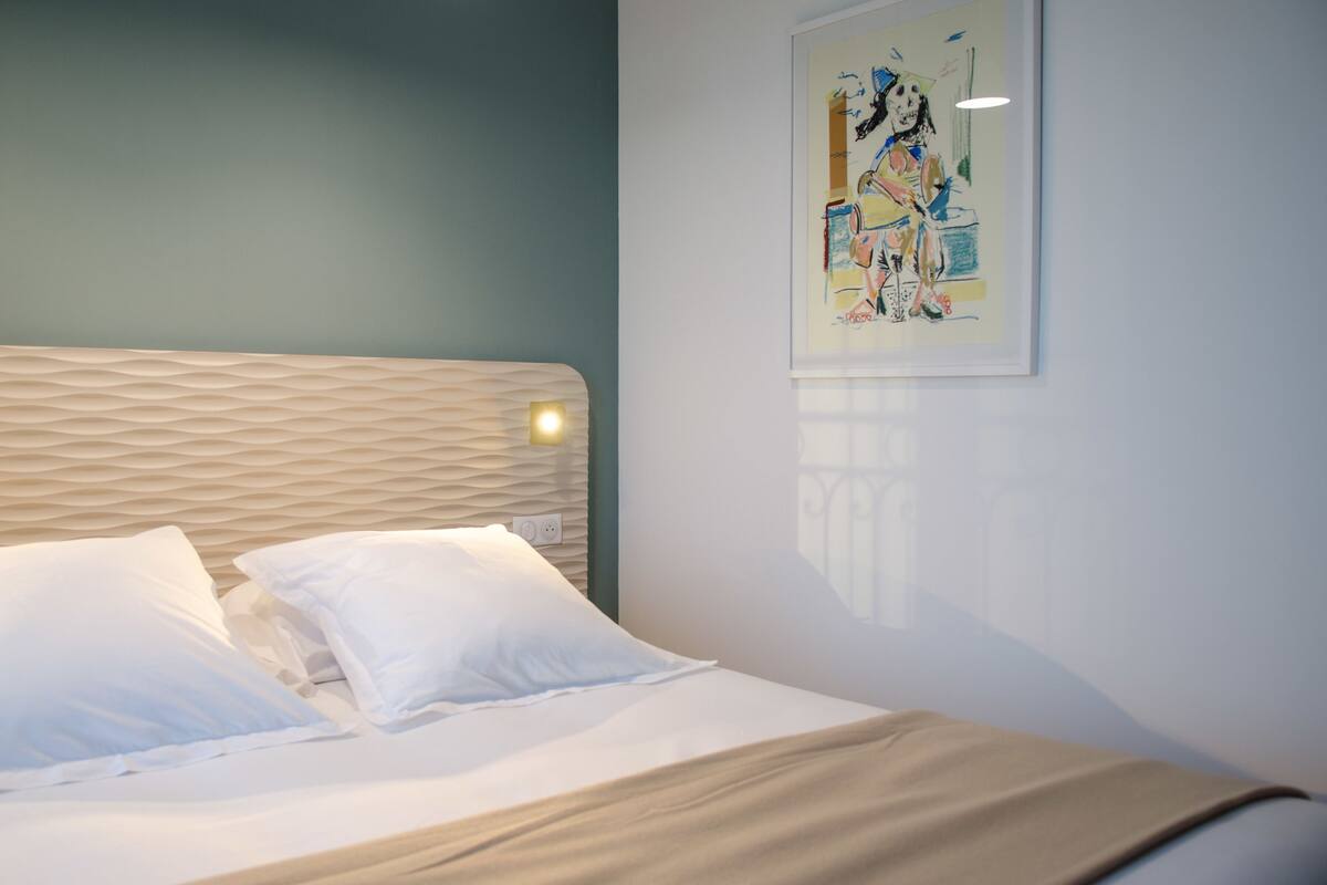 A neatly made bed with white linens and a beige headboard, a wall-mounted light, and a framed abstract painting.
