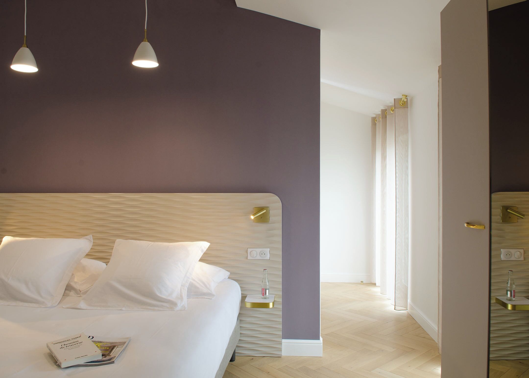 A modern hotel room with a large bed, white linens, and a magazine on the bed. There are two pendant lights above the bed and a wooden headboard.