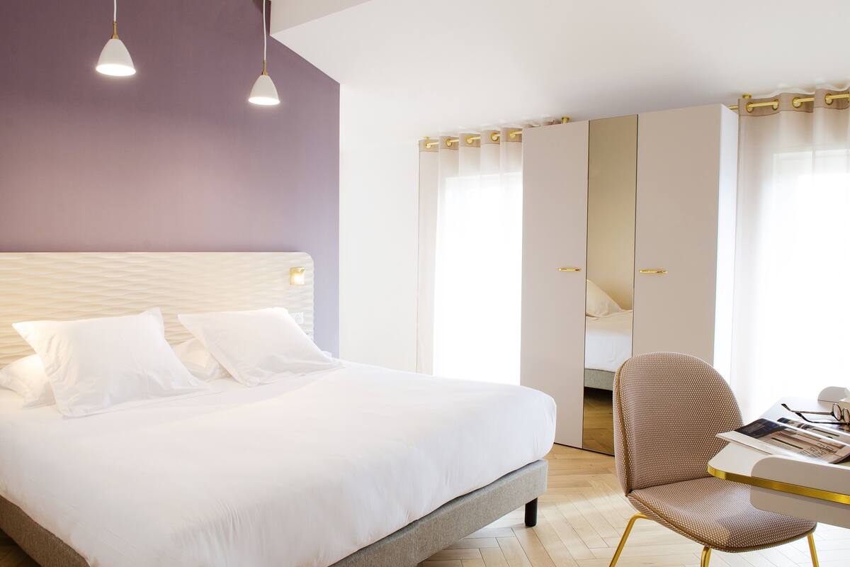 A modern hotel room with a large bed, a desk with a chair, and a wardrobe with a mirror.