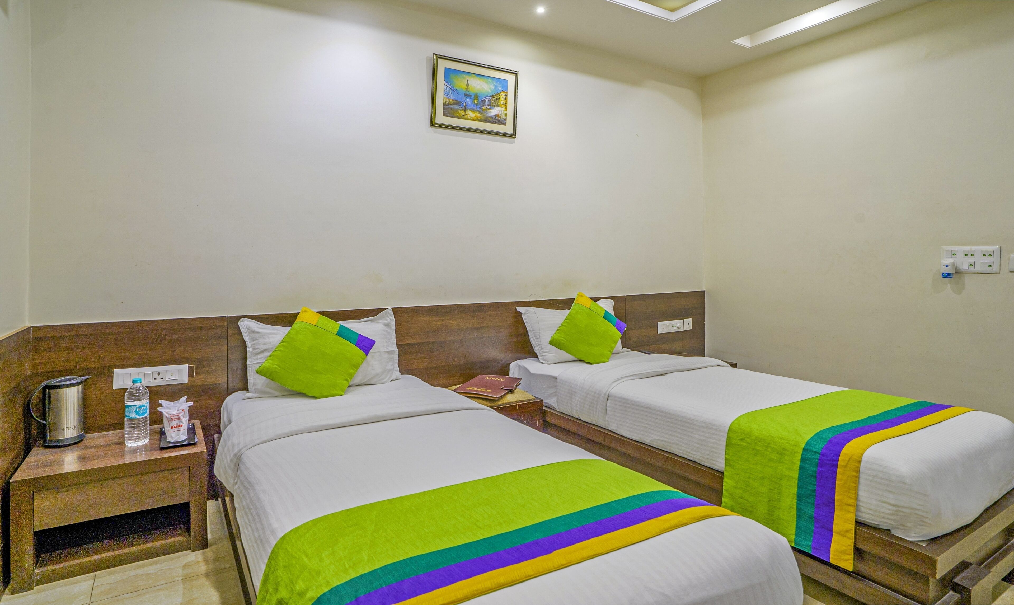 A hotel room with two beds, each with a green and purple striped blanket, a nightstand with a kettle and a bottle of water, and a painting on the wall.