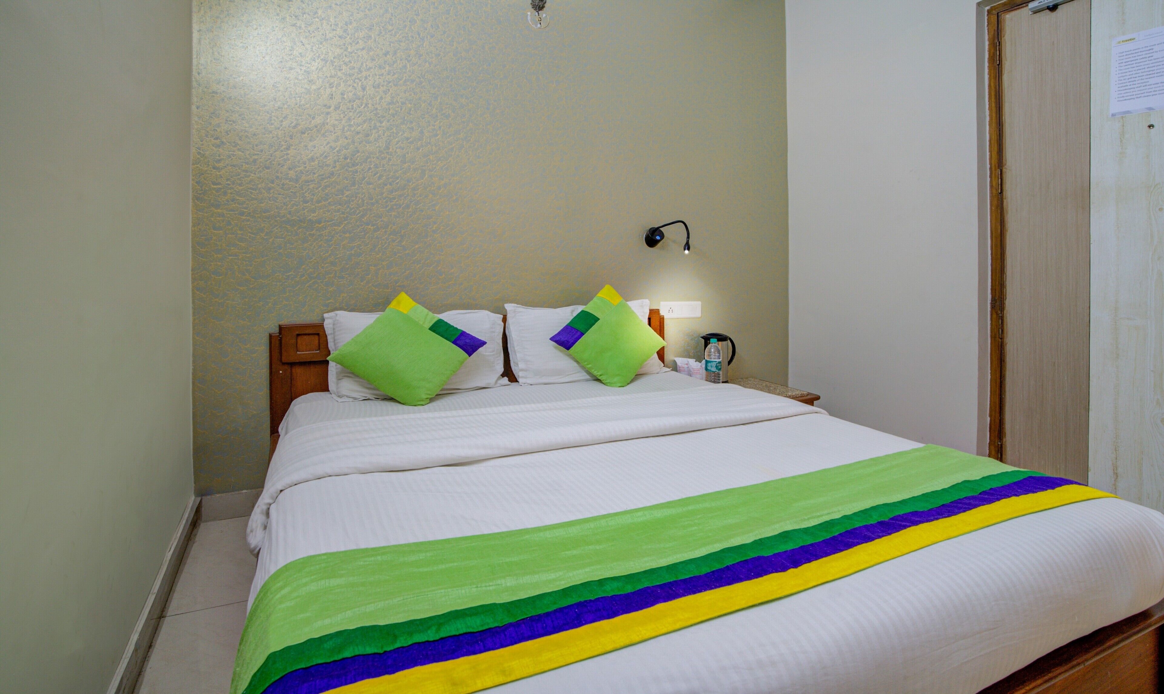 Una camera d'albergo con un letto, cuscini, un comodino, una lampada e una porta.