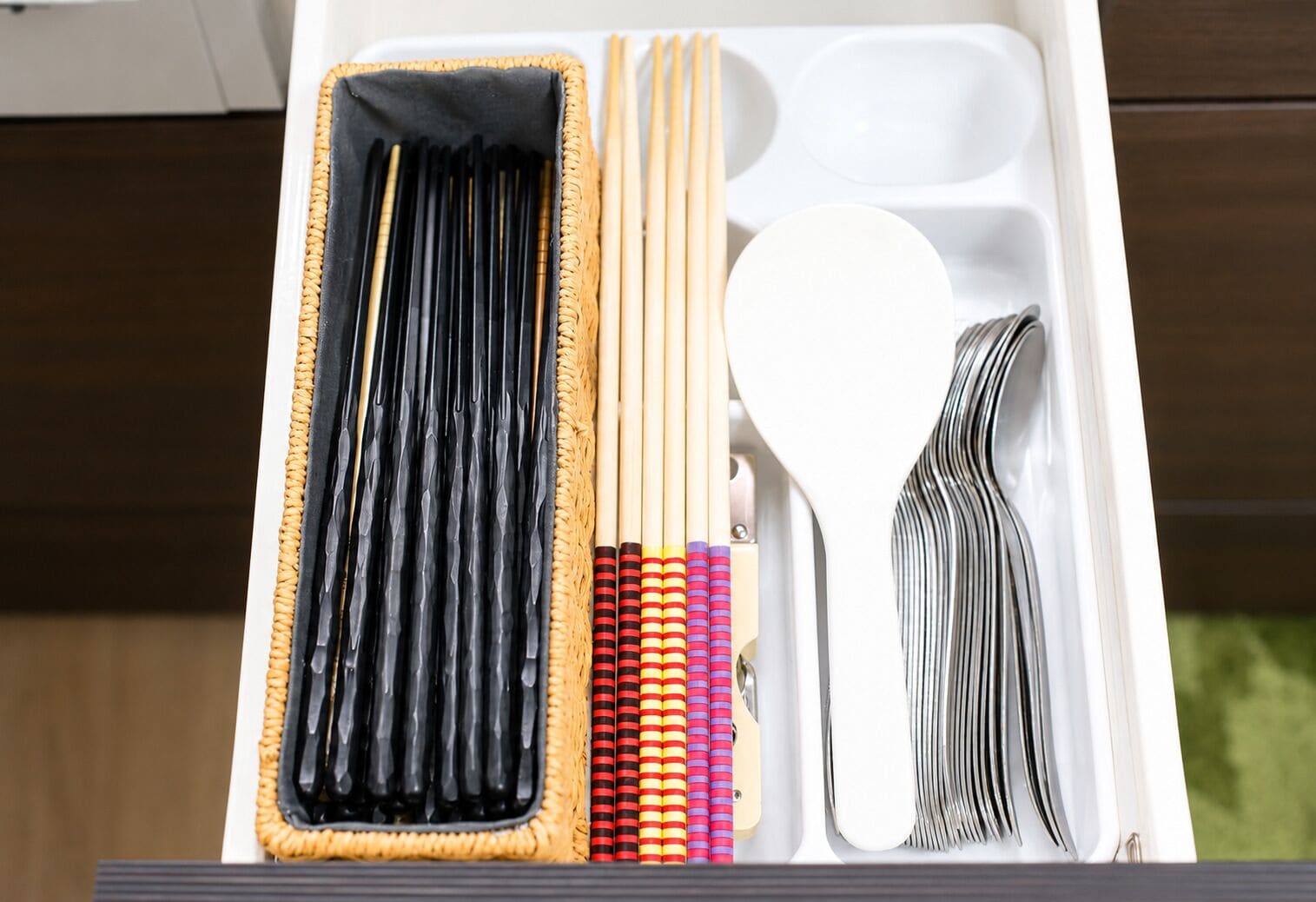 Un cajón con palillos negros, palillos de madera, una espátula blanca y un juego de palillos de colores con rayas.