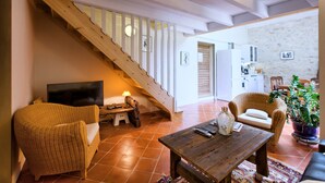 A living room with a wooden staircase, a TV, a sofa, a coffee table, and a kitchen in the background. - La Grange d'Oustaud (Verac)