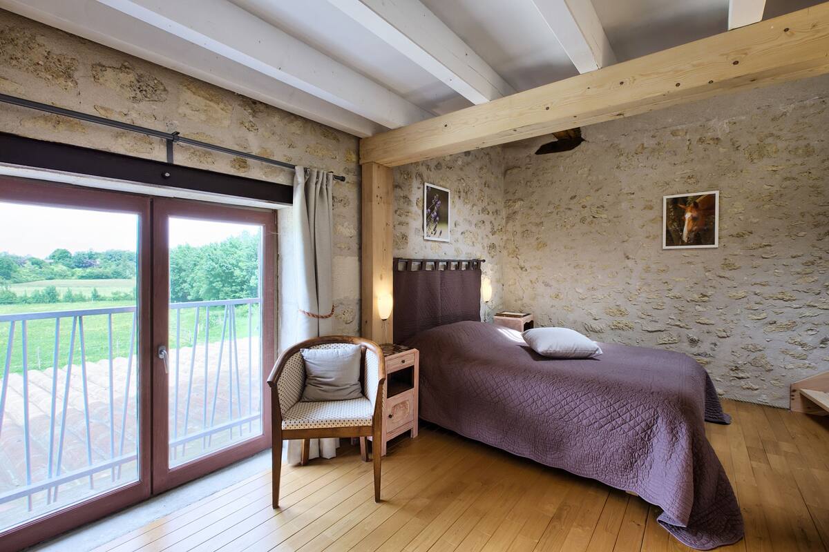 A bedroom with a large bed, a chair, a nightstand, a wooden ceiling, and a view of greenery through a sliding door.