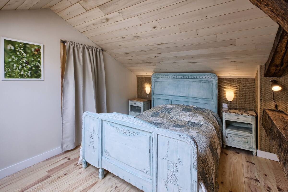 A bedroom with a wooden ceiling, a bed with a patterned headboard, two bedside tables with lamps, and a framed picture on the wall.