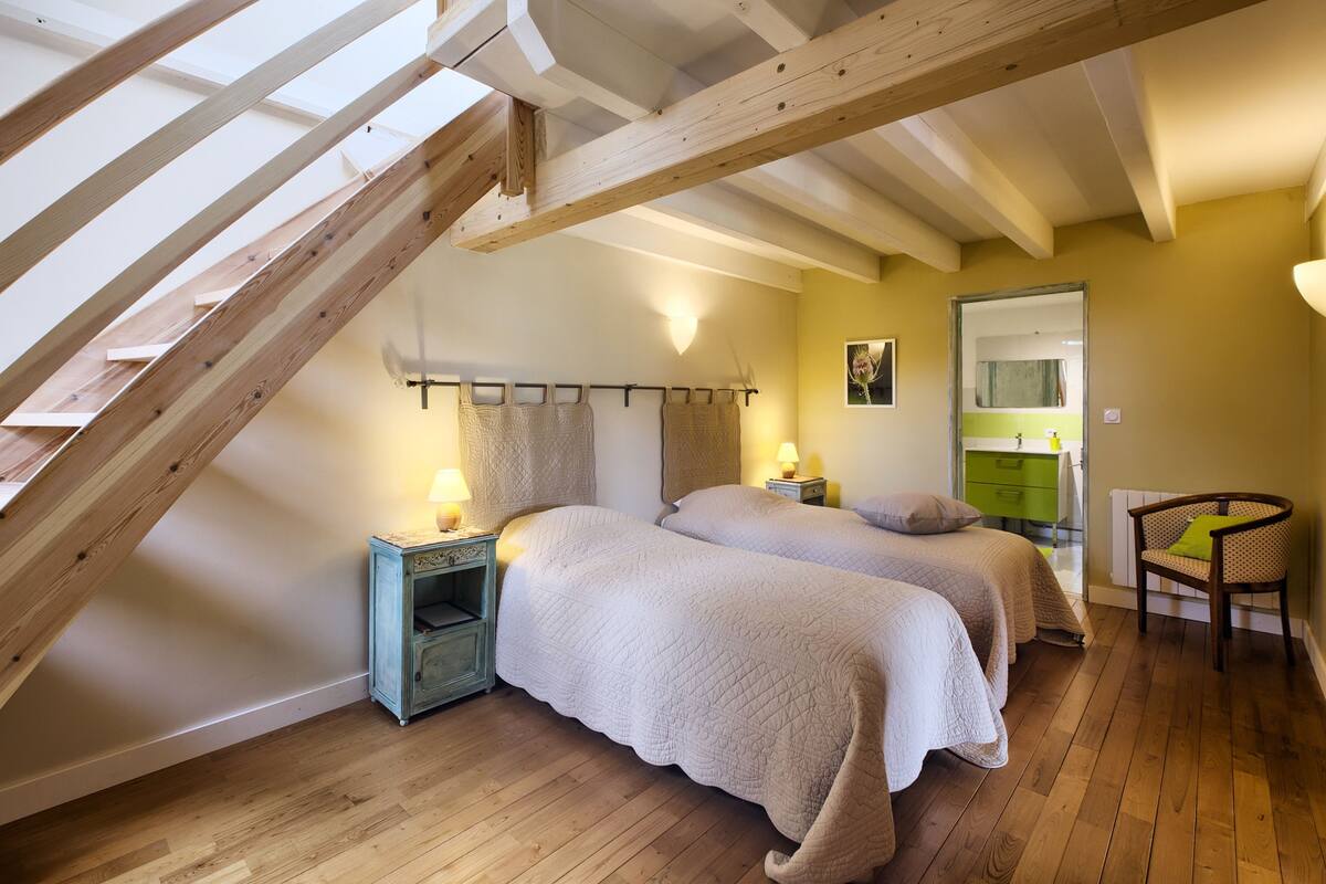 A bedroom with two beds, wooden beams, a skylight, a nightstand, a chair, and a bathroom visible through an open door.