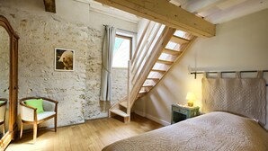 A bedroom with a sloped ceiling, a wooden chair with a green cushion, a bed with a quilt, a small bedside table with a lamp, and a picture on the wall. - La Grange d'Oustaud (Verac)