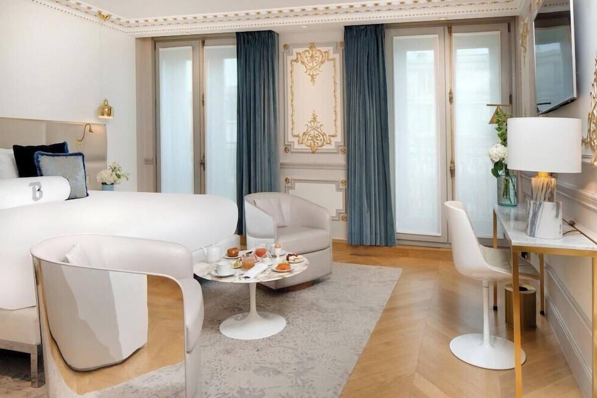 A modern hotel room with a large bed, a sitting area with a table, and a desk with a lamp.