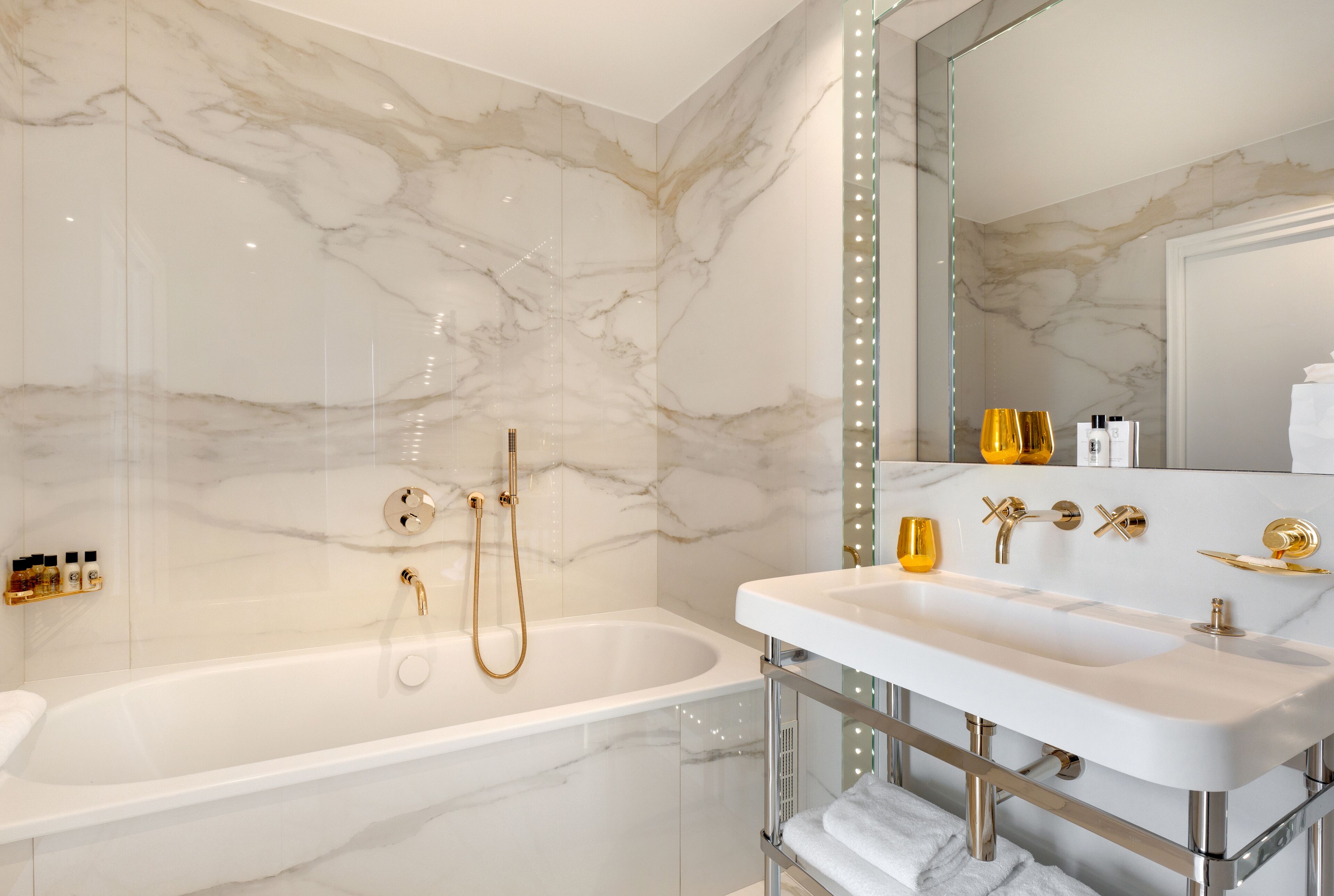 A modern bathroom with a marble wall, a bathtub, a vanity with a sink, and a mirror with lights.