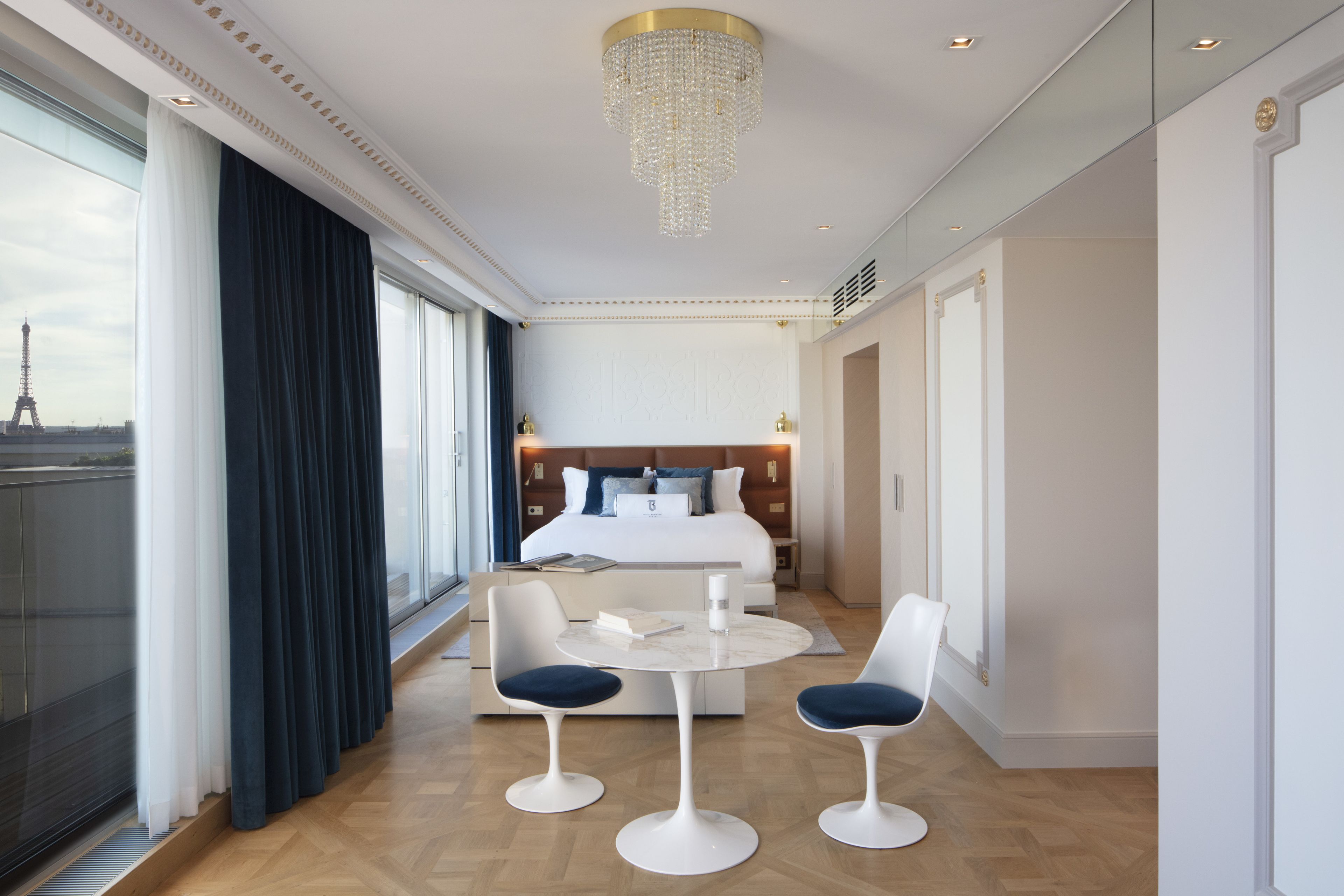 A modern hotel room with a large bed, a dining area with two chairs and a table, and a view of the Eiffel Tower through large windows.