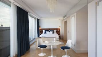 A modern hotel room with a large bed, a dining area with two chairs and a table, and a view of the Eiffel Tower through large windows.