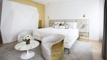 Un dormitorio moderno con una cama, una butaca blanca curvada, una mesita redonda con un jarrón de flores y una mesita de noche con una lámpara.