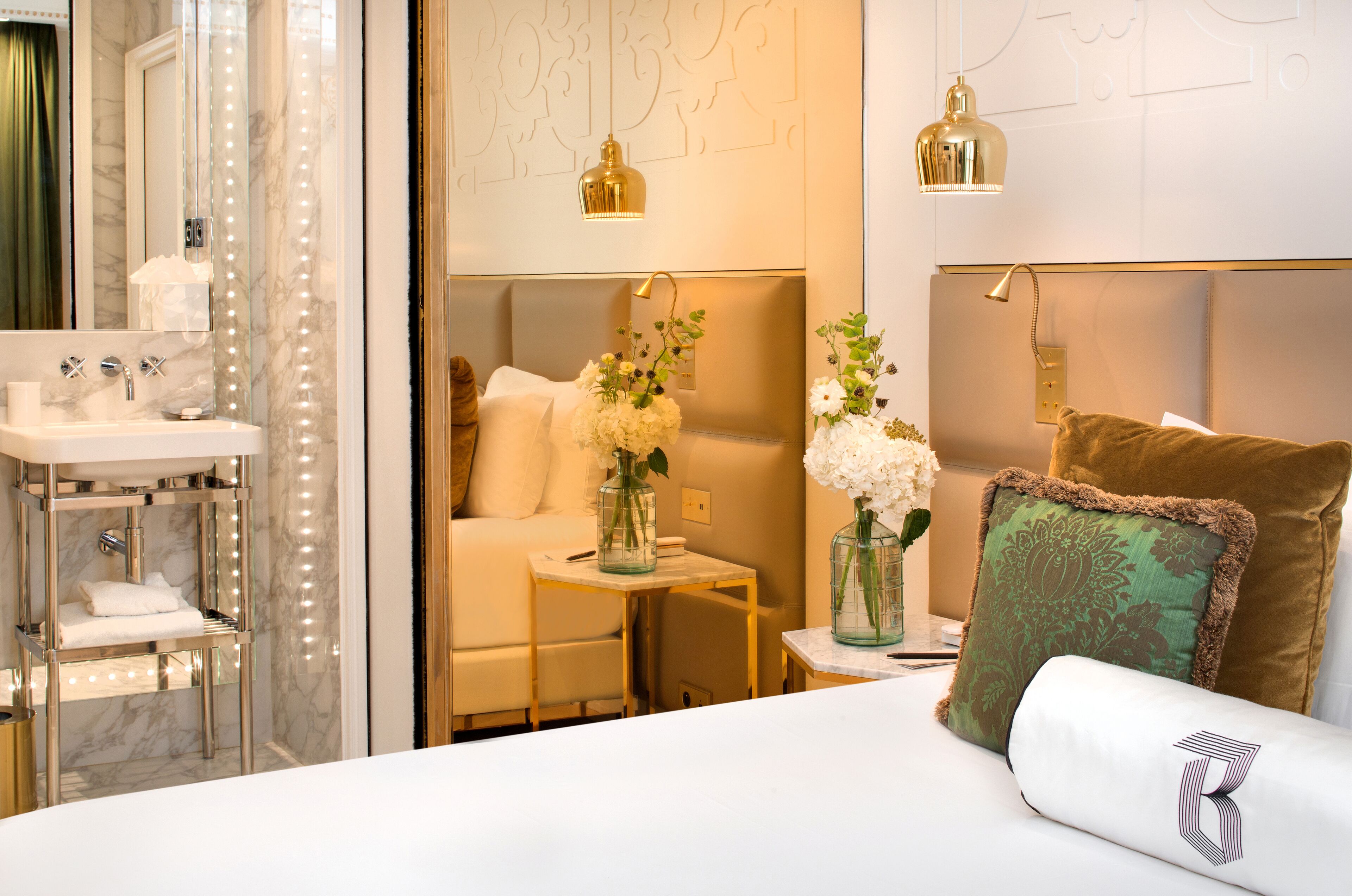 A hotel room with a bathroom, a bed with white linens, a small table with a vase of flowers, and a wall-mounted lamp.
