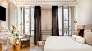 A luxurious bedroom with a large bed, a seating area with a white sofa, a gold table, and a view of the city through large windows. - Hôtel Bowmann, a Small Luxury Hotel of the World (Paris)