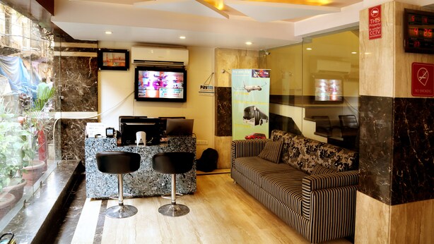 A hotel reception area with a marble counter, two bar stools, a sofa, and a television displaying an advertisement. - Hotel Geetasaar (New Delhi)
