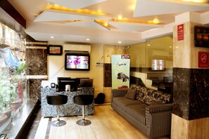 A hotel reception area with a marble counter, two bar stools, a sofa, and a television displaying an advertisement. - Hotel Geetasaar (New Delhi)
