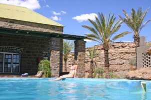 Zona de piscina con un edificio de piedra, palmeras y cielo despejado.