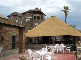 Un patio con sillas y mesas de plástico blanco, una estructura de techo de paja y un edificio de piedra al fondo.