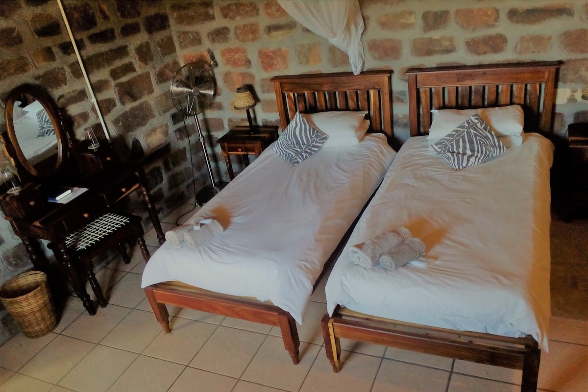 A room with two wooden beds, a small desk, a chair, a mirror, and a stone wall.
