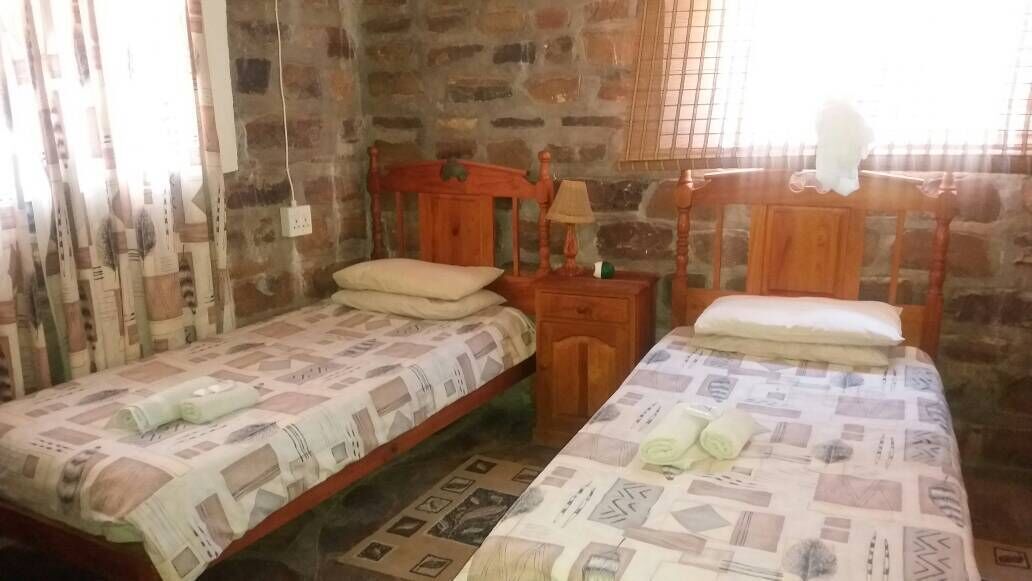 A room with two single beds, a wooden headboard, a nightstand with a lamp, and a stone wall.