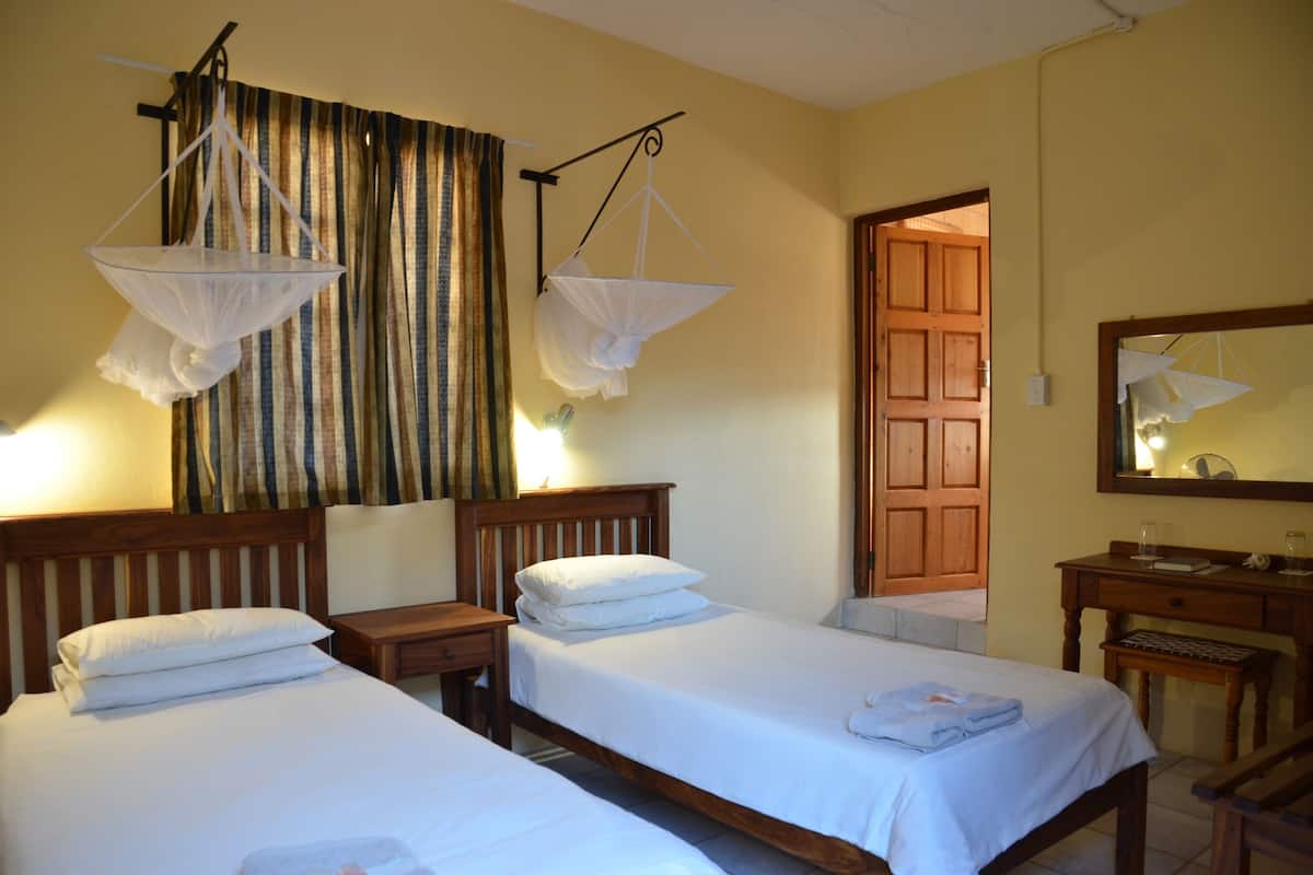 A room with two single beds, wooden headboards, a wooden desk, a chair, a mirror, and a door leading to another room.