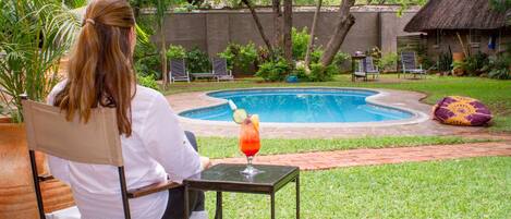 A person seated at a table with a drink, overlooking a swimming pool surrounded by greenery and a thatched-roof structure.