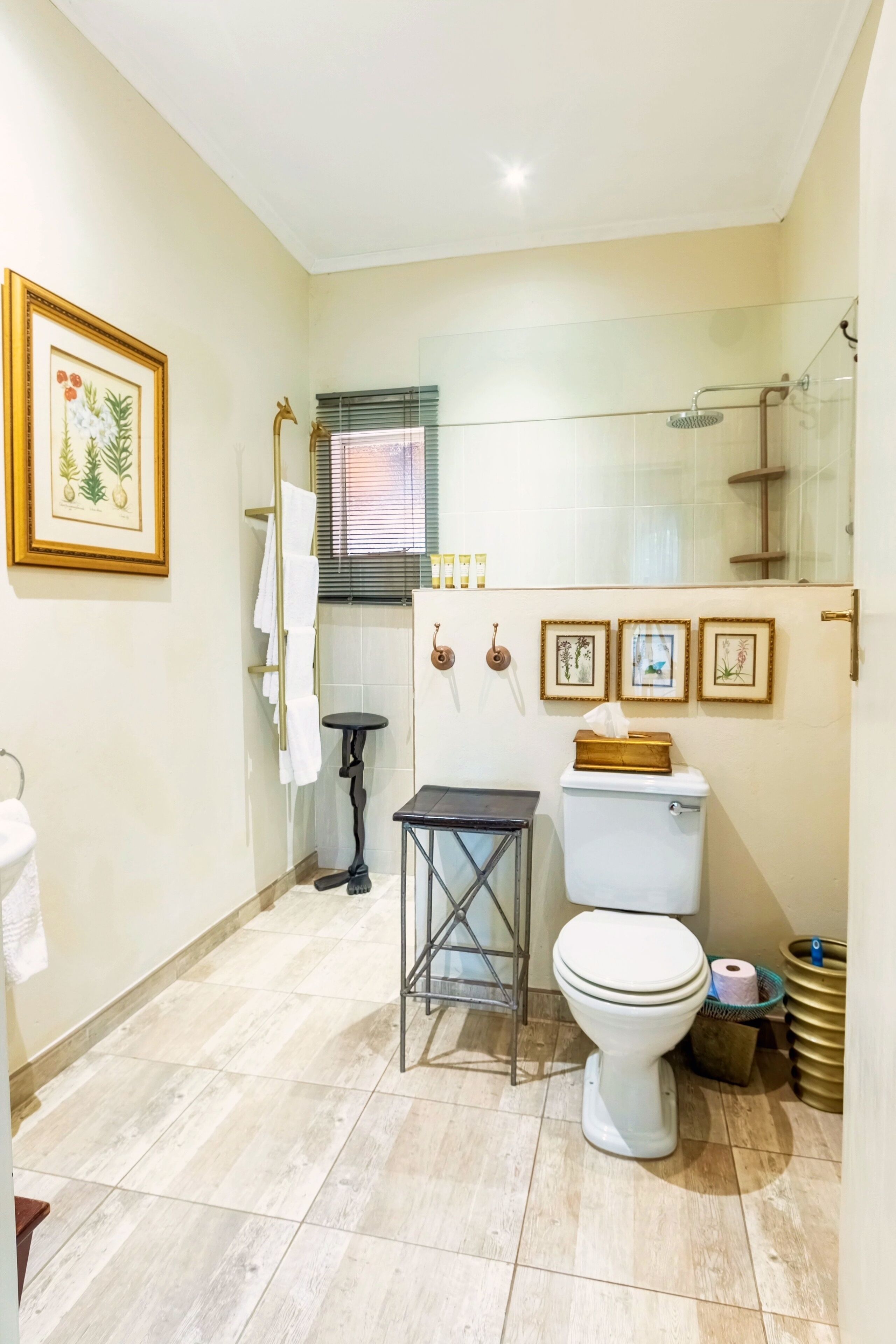 Un baño con inodoro, lavamanos y ducha. Hay cuadros enmarcados en la pared y una ventana con persianas.