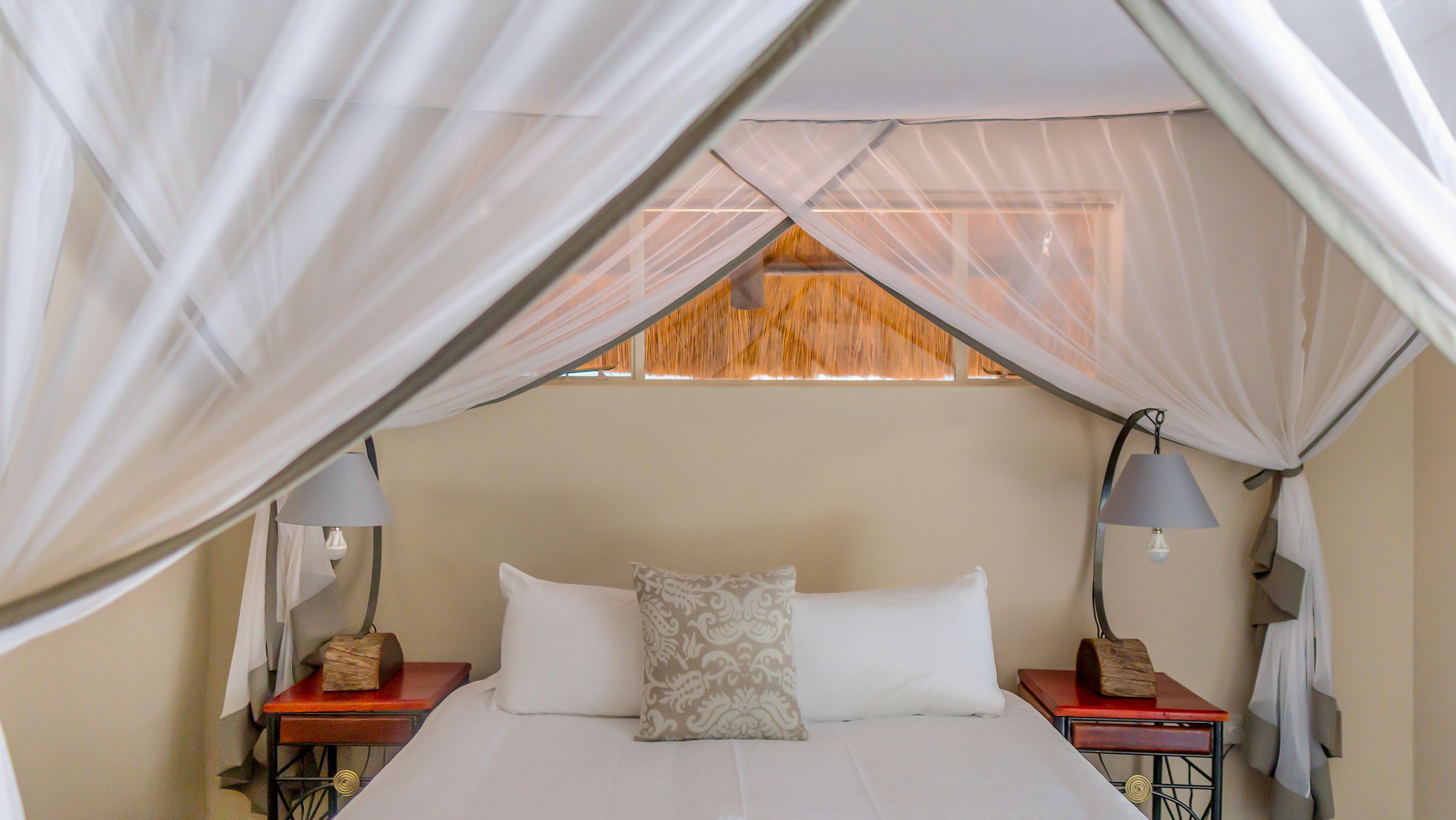 A bed with white linens, a patterned pillow, and a canopy overhead. There are two bedside tables with lamps and a small wooden block on each.