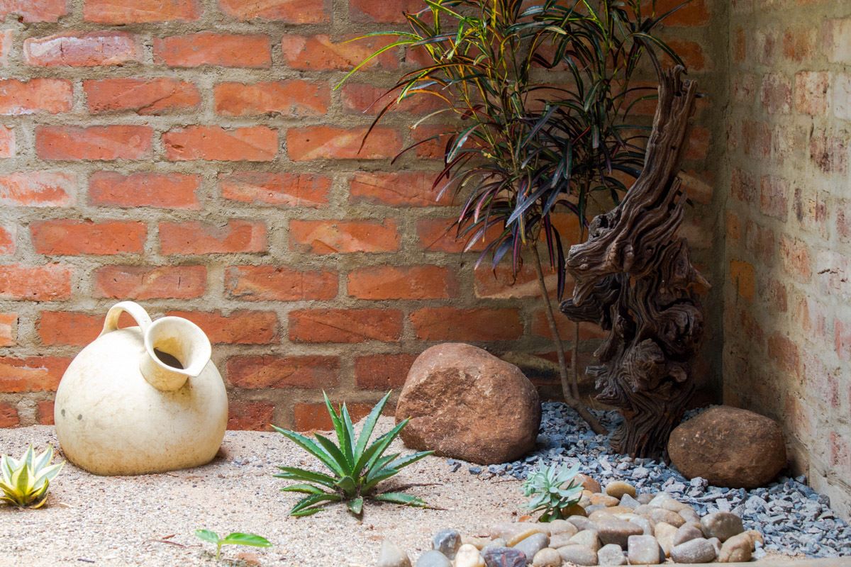 Um ambiente rústico ao ar livre, com uma parede de tijolos, um tronco de árvore decorativo, um jarro de cerâmica e diversas pedras e plantas.