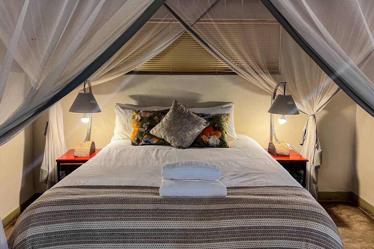 A neatly made bed with a striped blanket, a floral pillow, and a folded towel on top. There are two bedside tables with lamps and a canopy over the bed.