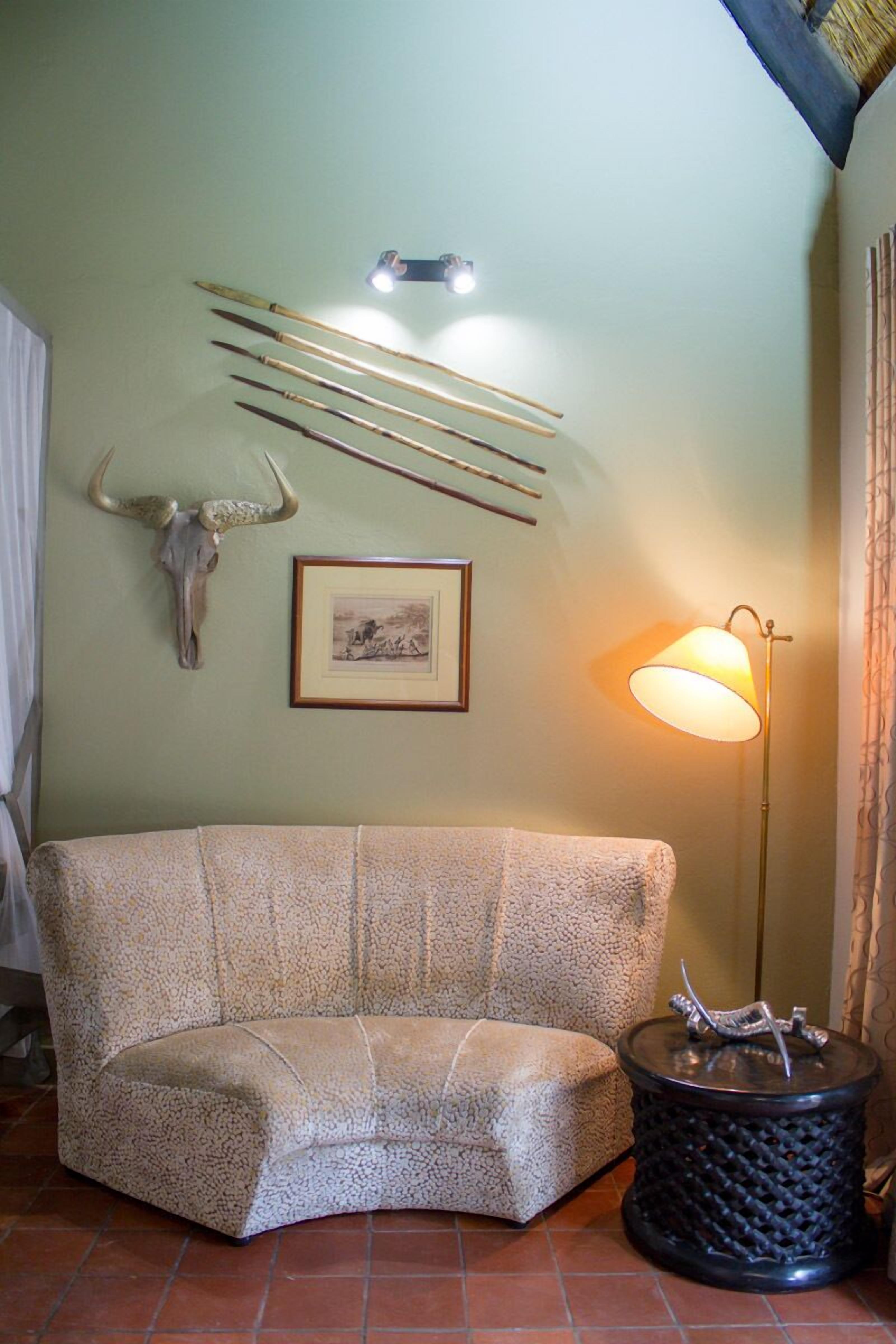 Een kamer met een gestructureerde bank, een aan de muur bevestigd herdershoofd, een ingelijst beeld, een vloerlamp en een klein rond tafeltje met een decoratief object.
