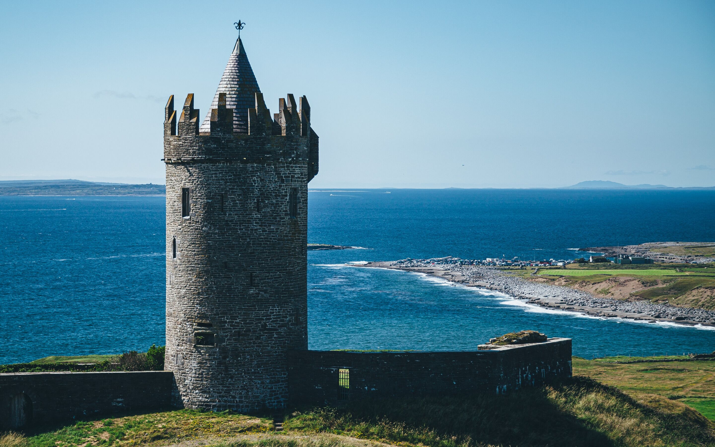A stone tower with a pointed roof and crenellations, overlooking a coastal area with a clear view of the sea and distant landforms.