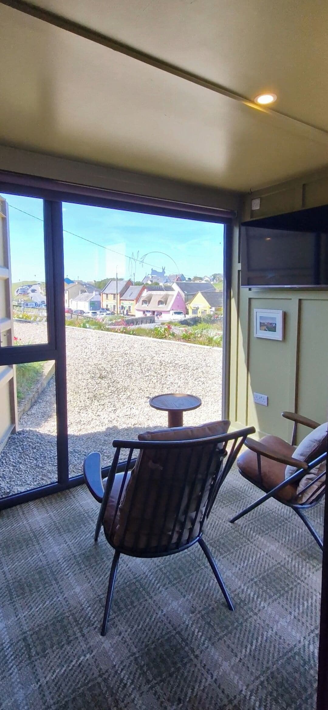 A room with a large sliding glass door leading to a gravel area with houses in the background. There is a small round table and two chairs with cushions.