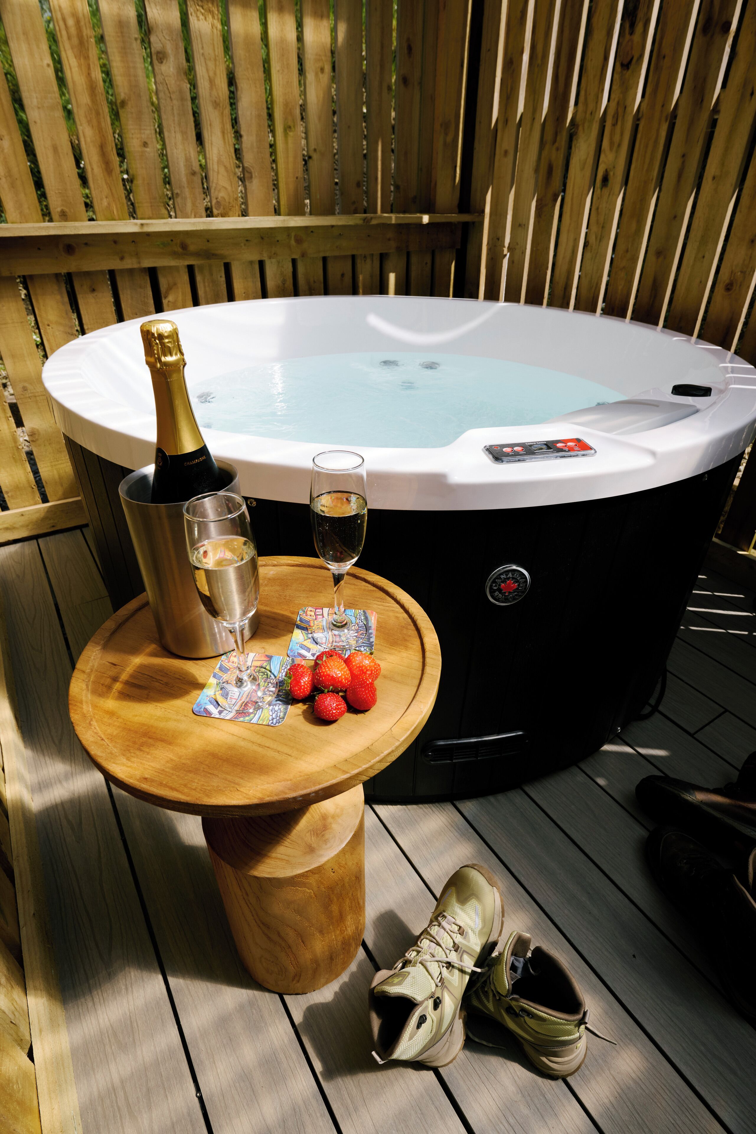 A hot tub with a champagne bottle and glasses, strawberries, and a wooden table on a wooden deck.