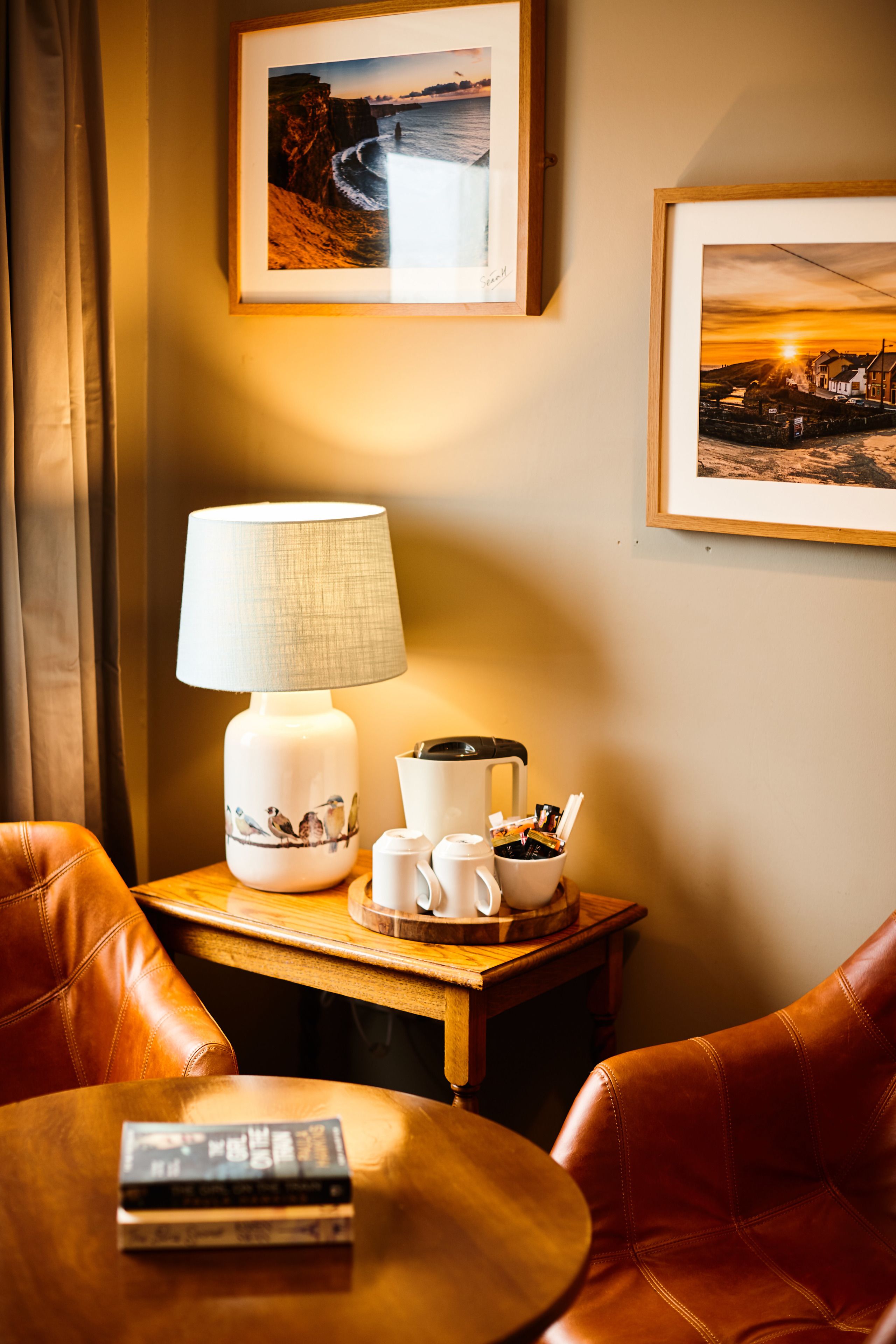 Una habitación de hotel con una mesita de madera, una lámpara, una cafetera y cuadros en la pared.