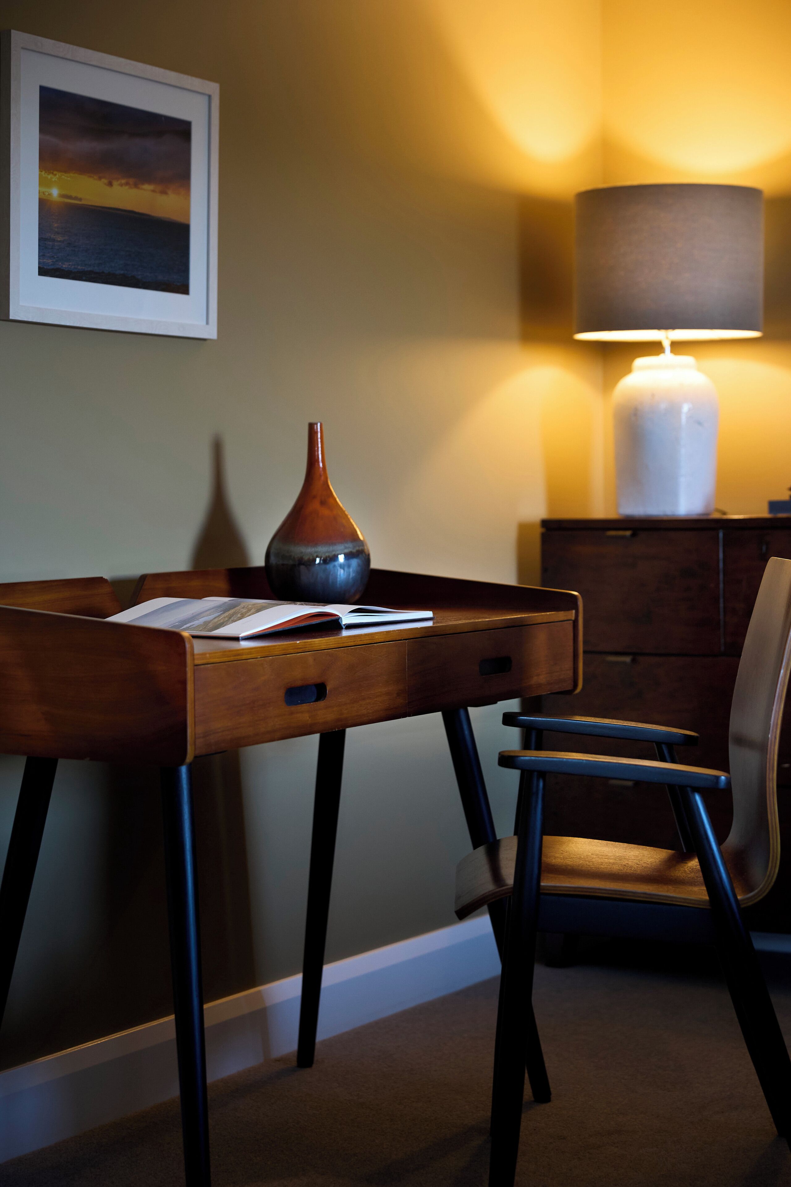 A wooden desk with a vase, a lamp, and a chair in a room with a painting on the wall.