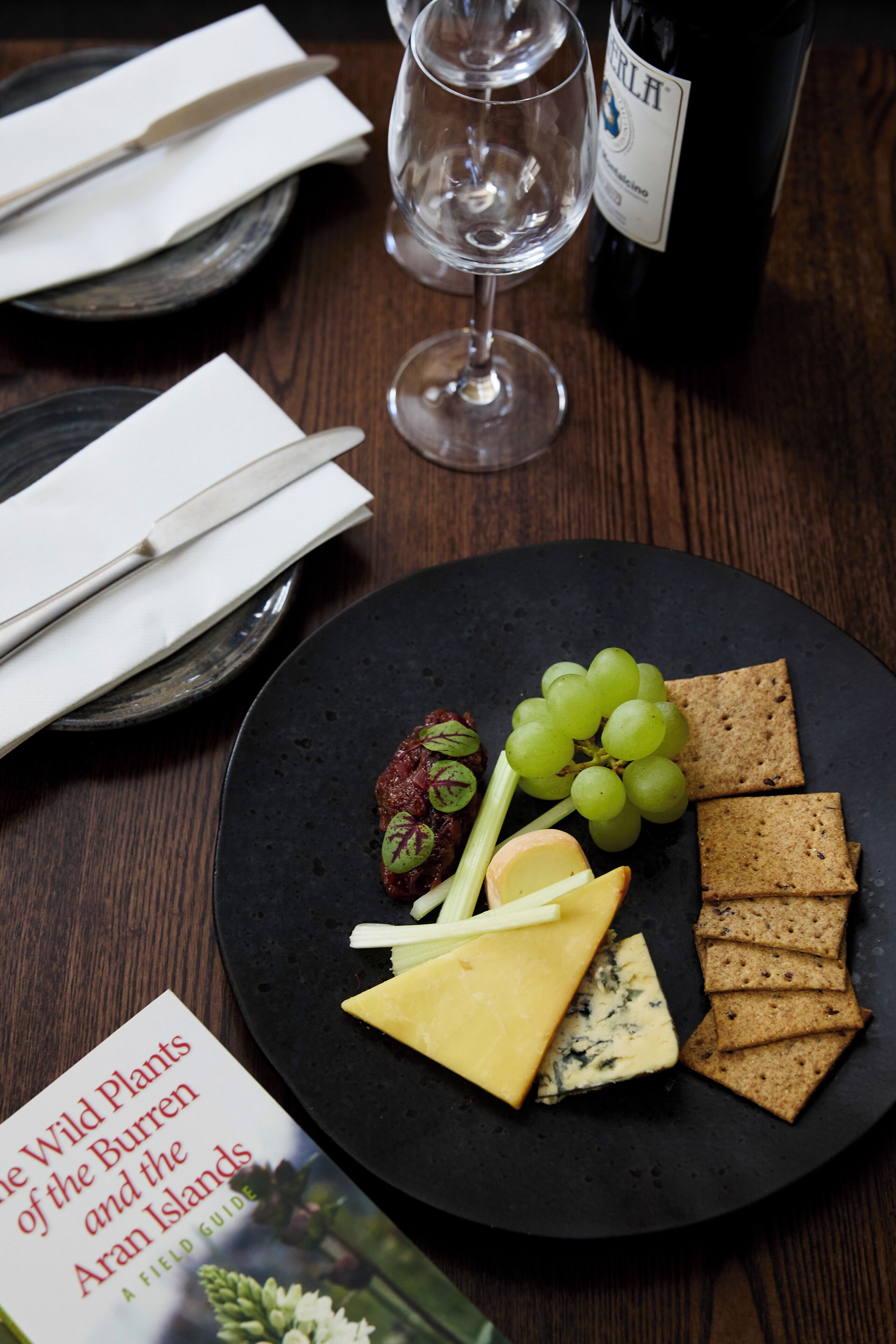 A plate with cheese, grapes, crackers, and a book titled "The Wild Plants of the Burren and the Aran Islands" on a wooden table.